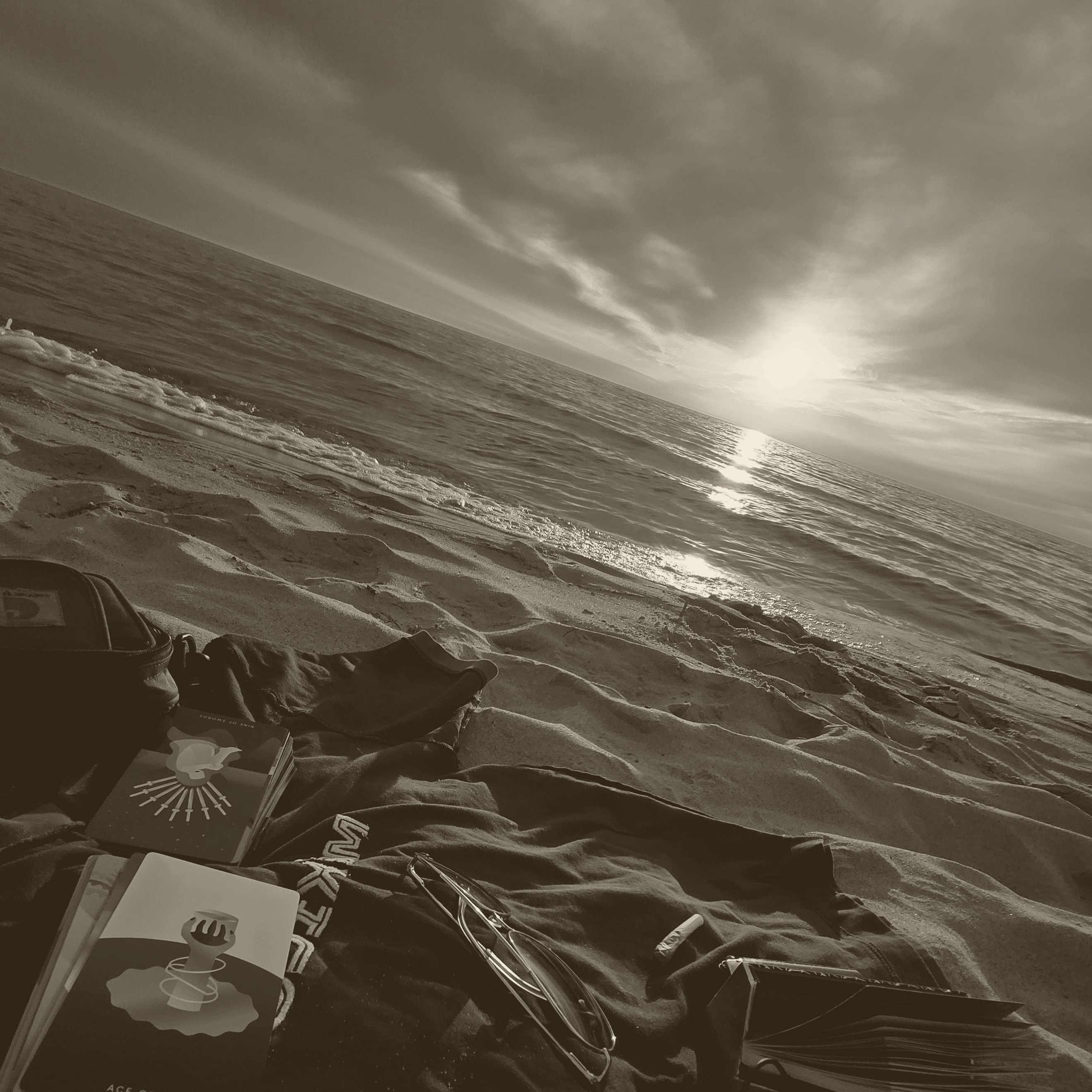 Sepia photo of a beach at sunset: tarot cards, glasses, and a journal resting on a blanket in the sand beside a calm lake